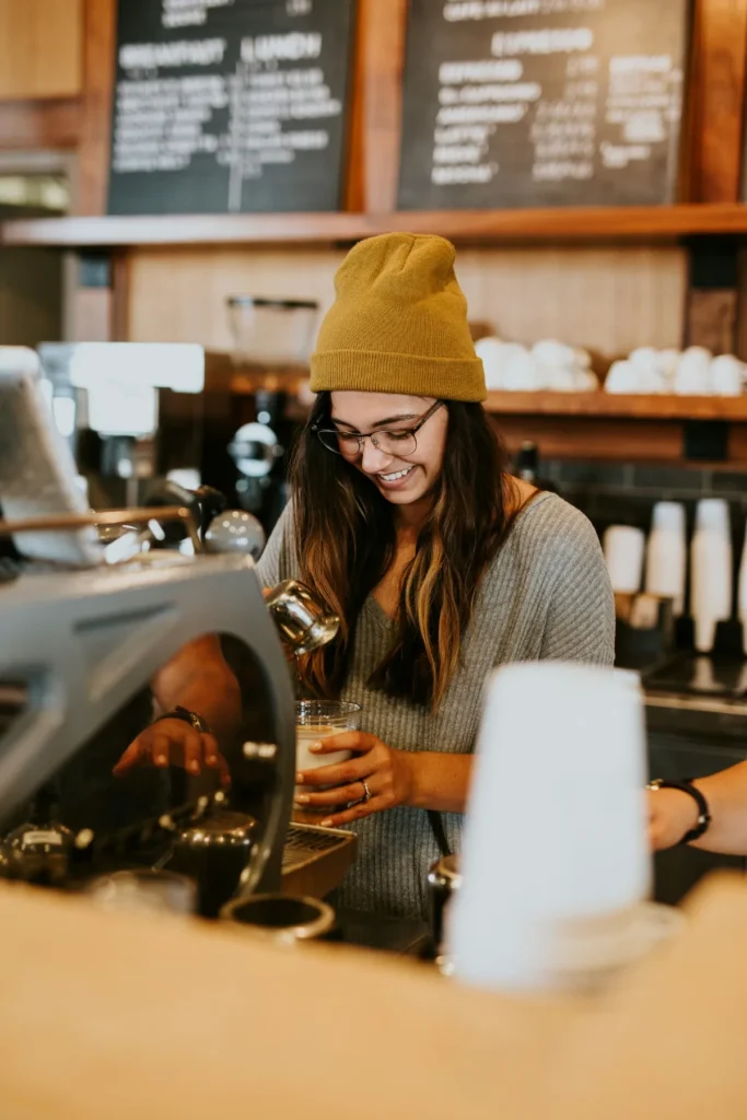 Barista sorridente preparando café espresso em cafeteria, de gorro mostarda e óculos.