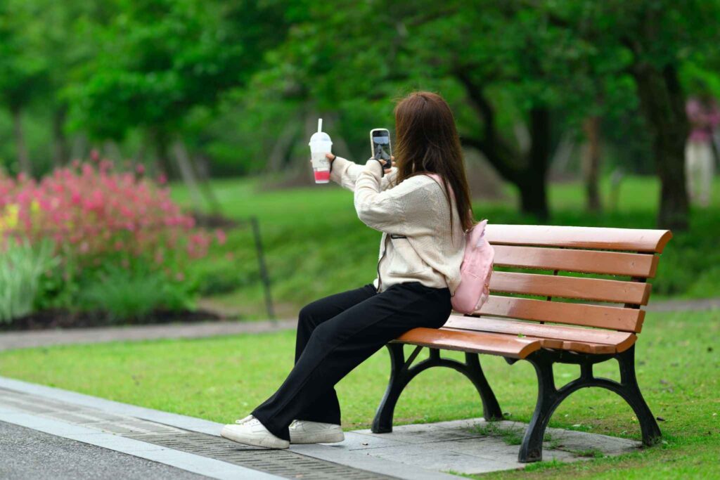 Jovem sentada em banco de parque segurando celular e bebida, registrando uma selfie.